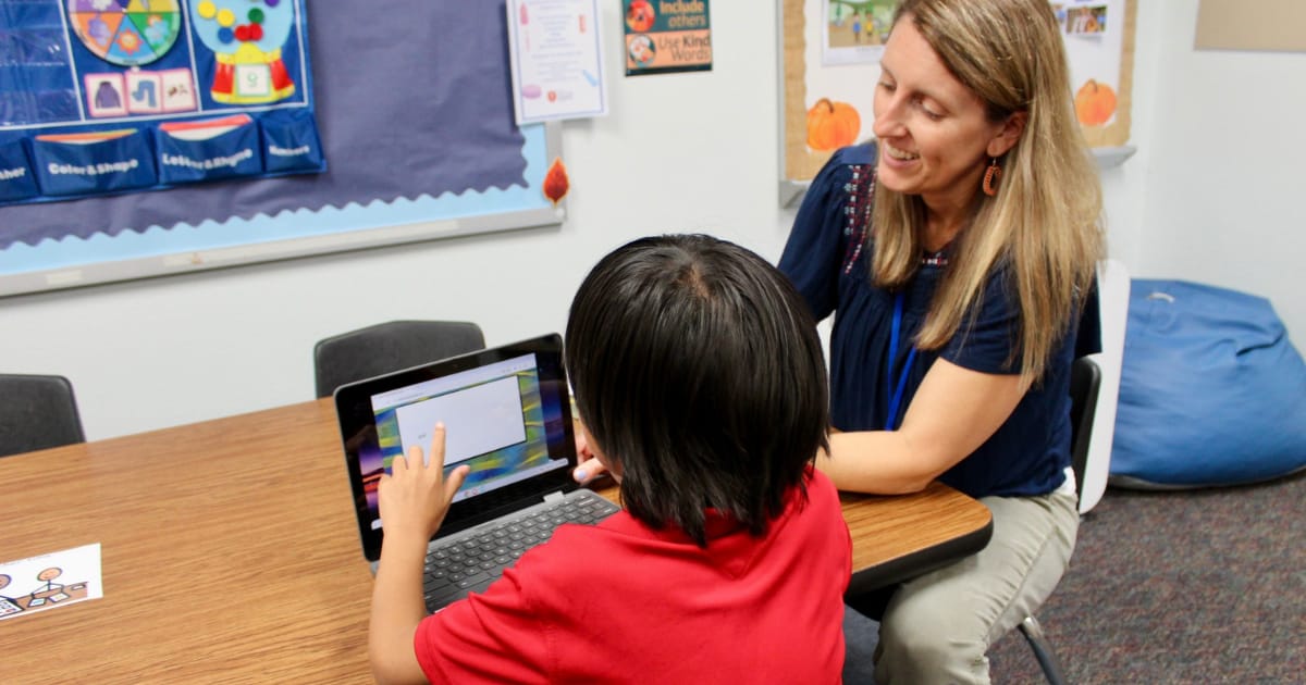 Tuttle teacher smiles while looking at a student that is looking at an iPad.