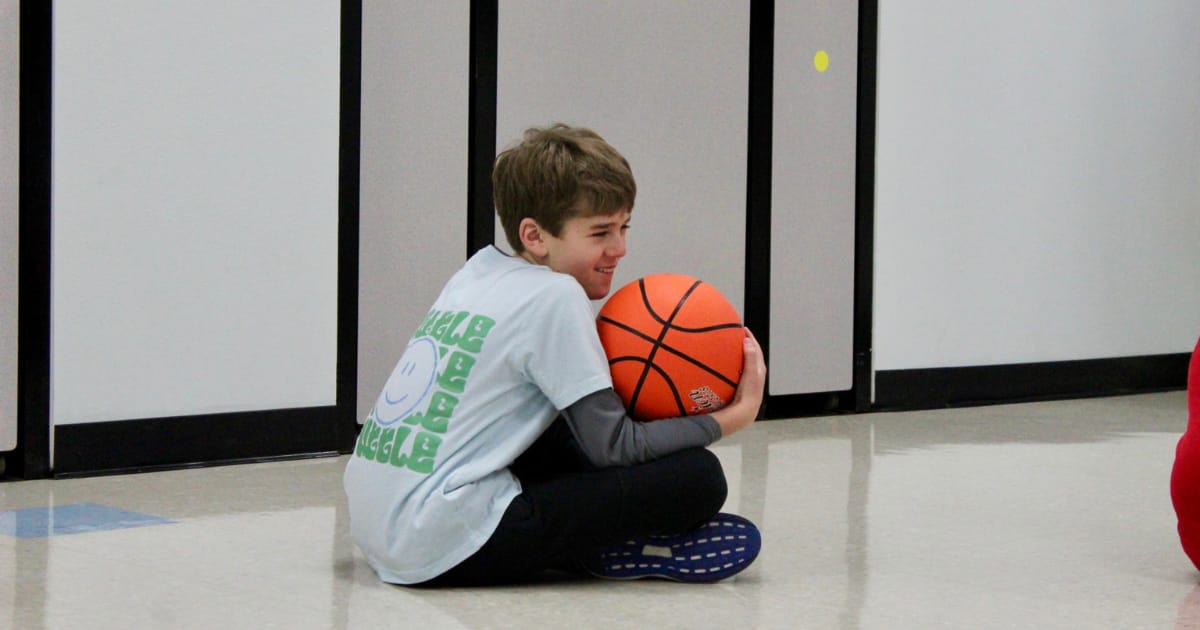 Tuttle student sits on the ground while smiling and holding a basketball in his lap.