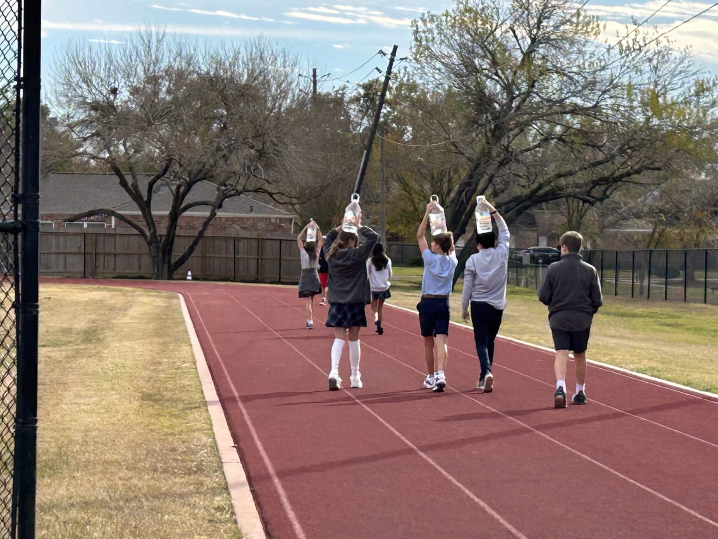 Students walk on a track with gallon jugs of water on their heads.