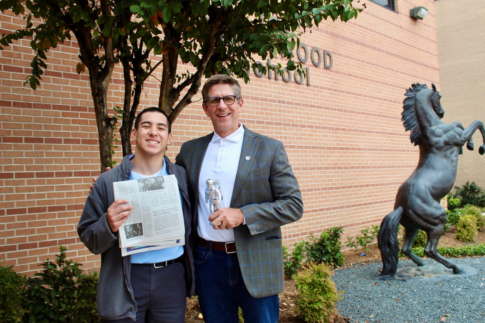 Student and head of school stand together outside of Briarwood building and smile. Student holds a newspaper, head of school holds an astronaut stature.